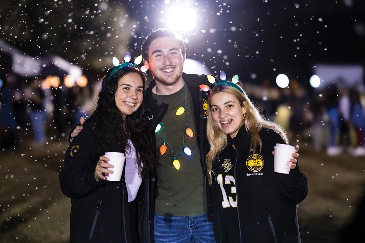 Three students pose for a photo while wearing Christmas light accessories as fake snow falls