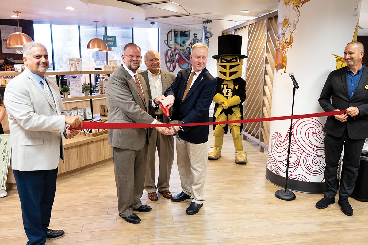 Two men hold a red ribbon while three men hold a large pair of scissors. 