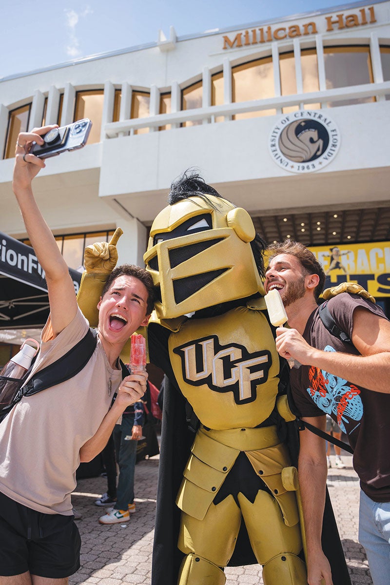 Students take a selfie with Knightro while holding popsicles in front of Millican Hall.