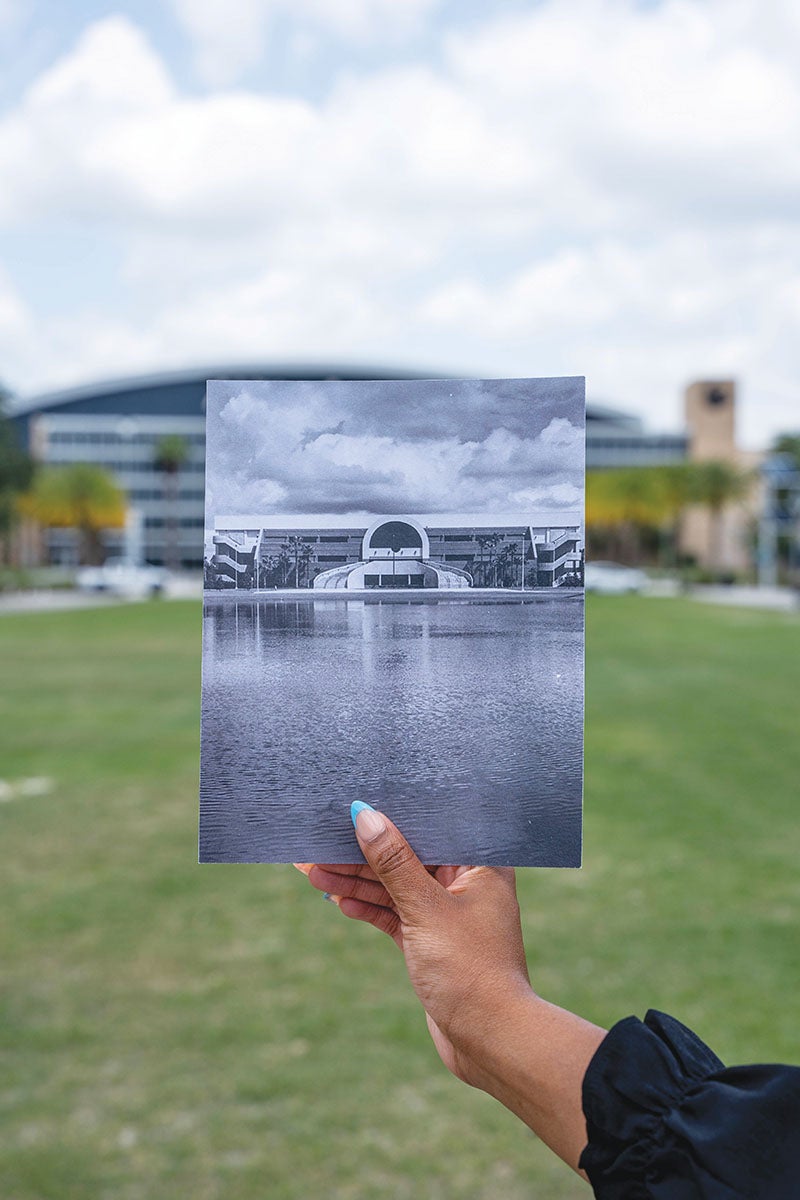 A person holding a photo of the Addition Financial Arena with the arena in the background behind the photo