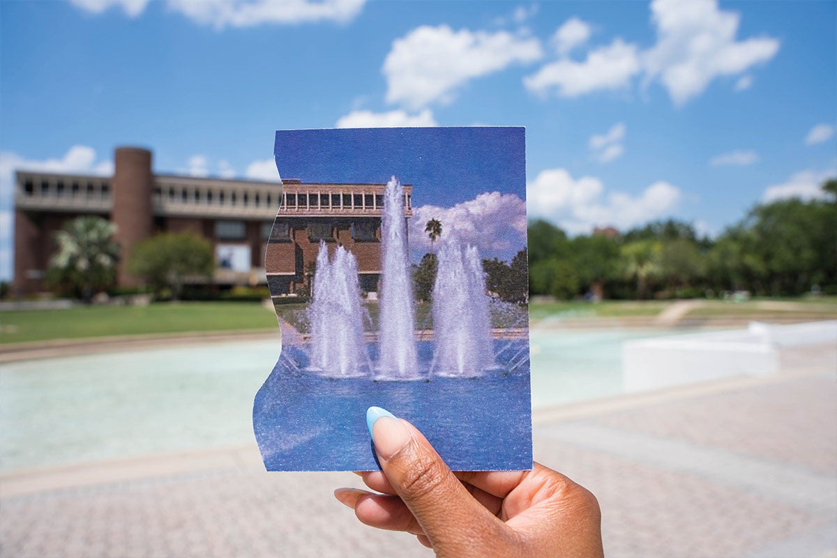 A hand holding a photo of the reflecting pool fountain in front of the John C. Hitt Library