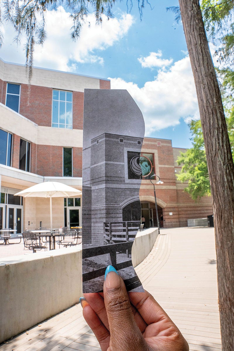 A hand holding a black and white photo of the Student Union building with the current union building behind the photo