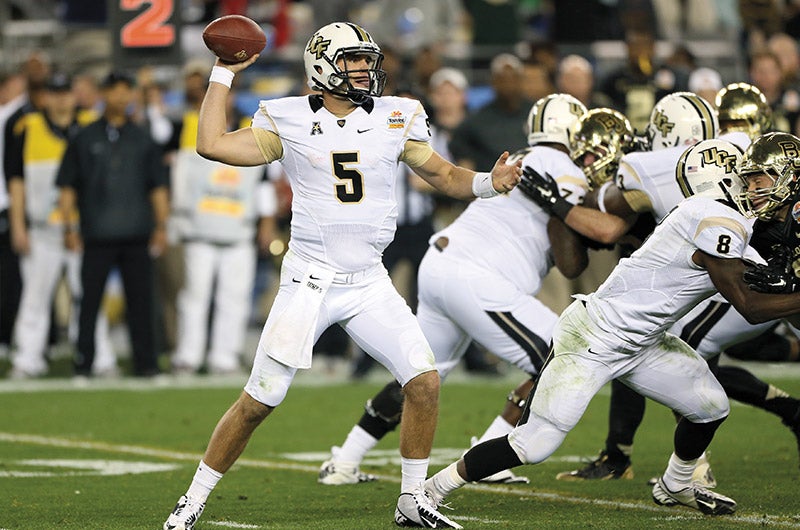 A man holds a football while his teammates hold off players from the opposing team.