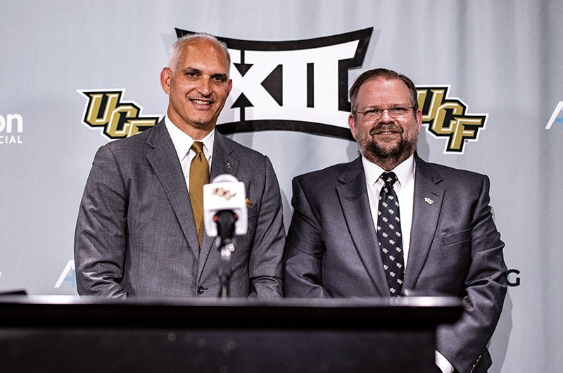 Two men in suits standing at a podium with a backdrop with UCF and Big 12 logos behind them.