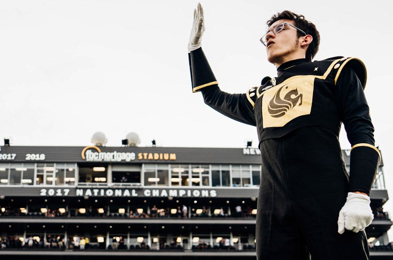 A marching band player holding his hand up while in the FBC Mortgage Stadium with the words "2017 National Champions" displayed