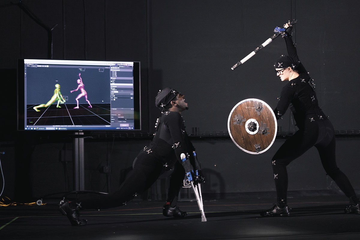 Two UCF students in full-body motion capture suits simulate a sword fight in the dark at UCF's Studio 500. Sensors track their movements, which are displayed as animated figures on a nearby screen. Instructors and equipment are visible in the background.