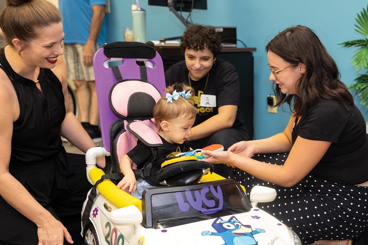 Child with blue bows in pigtails in her hair sits in child-sized white car decorated with Bluey on hood and the name Lucy in purple letters on black windshield. She is propped up with pink car seat insert and straps and purple kick board behind her back. Three adult women kneel down next to the car.