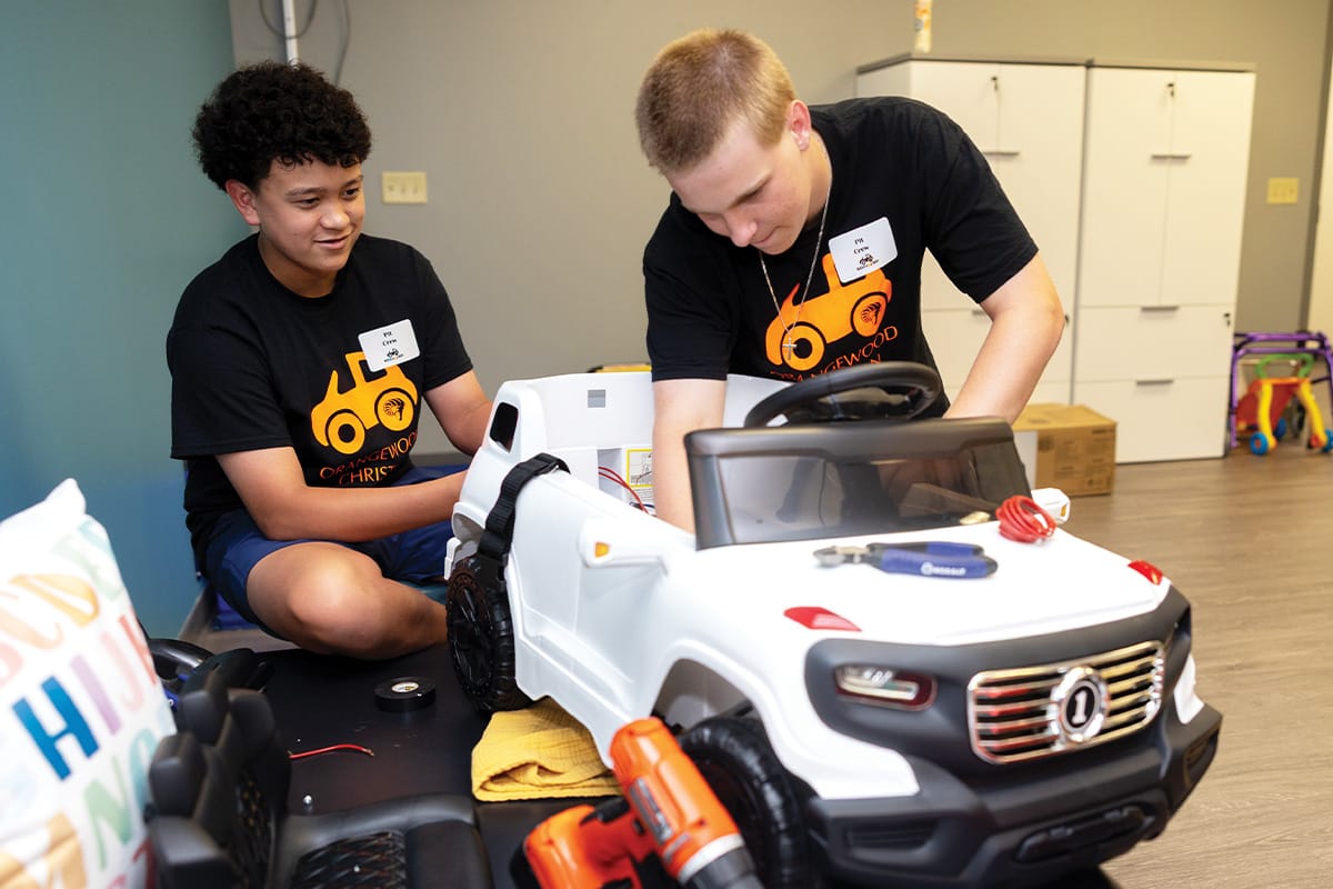 Two male students in black T shirts with orange car graphic on front work on adjusting white child-size car.