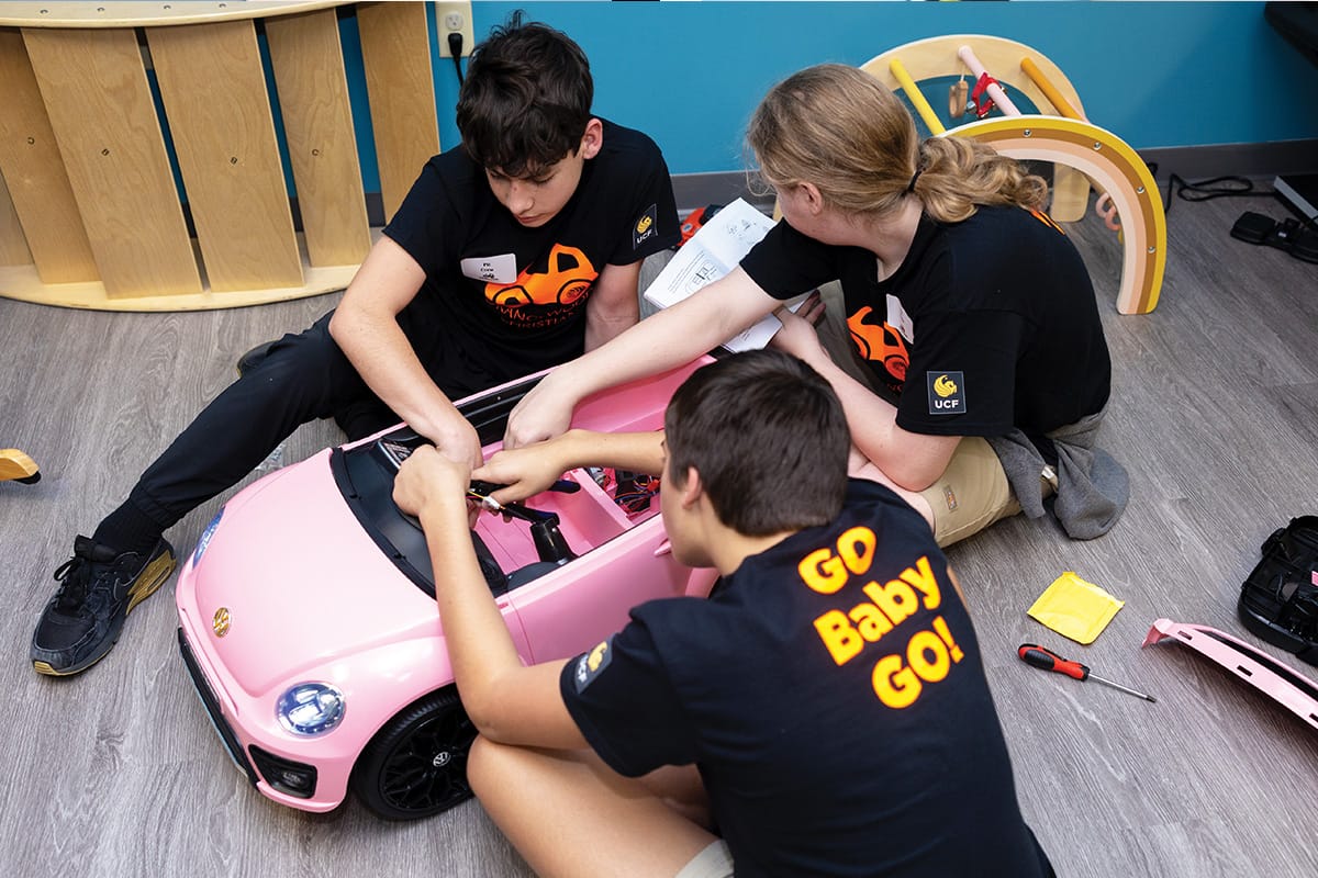 Three students wearing black shirts with orange car graphic on front and yellow Go Baby Go written on back tinker with pink car as they kneel on vinyl flooring.