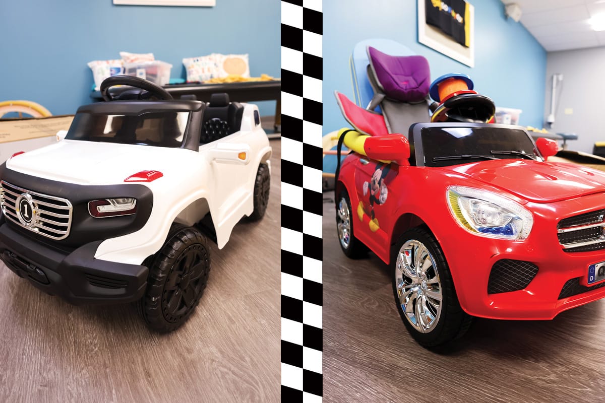 White and black child-sized SUV with silver grill and black wheels is parked on brown vinyl flooring in room with blue walls. On the right, a red child-sized car with Mickey Mouse sticker on passenger-side door is parked on brown vinyl flooring with blue walls.