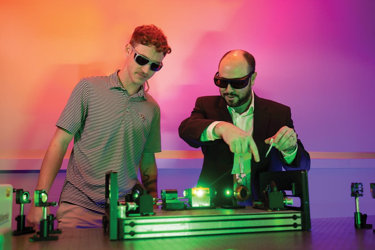 A researcher working with a student in an optics lab with colorful lights behind them