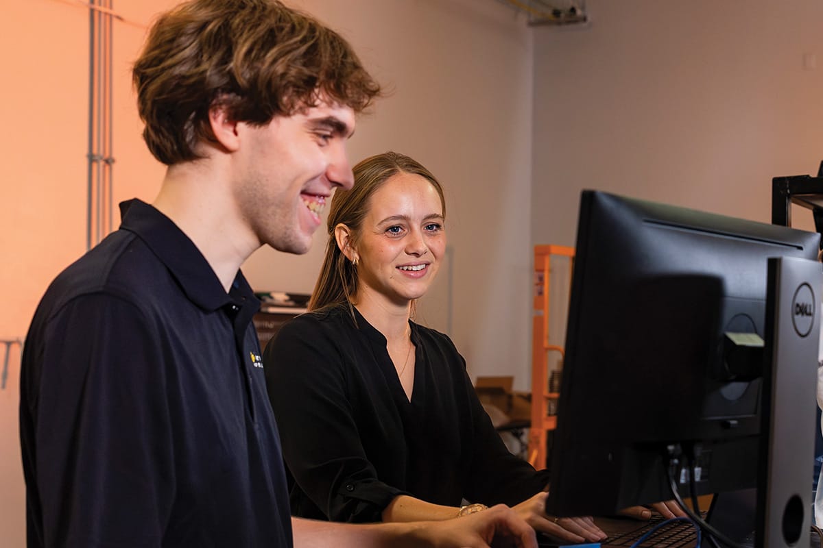 Side view of two UCF students standing at a computer workstation, smiling at the screen.