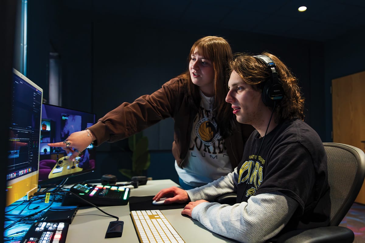 Woman wearing brown coat over white T shirt points to computer monitor as she stands next to man with black headphones seated at desk