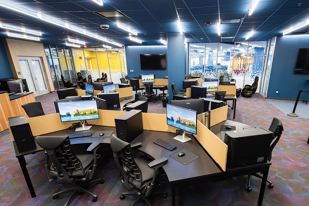 Wide angle of room with blue walls and empty desk stations equipped with computer monitor, keyboard, mouse and desk chairs.
