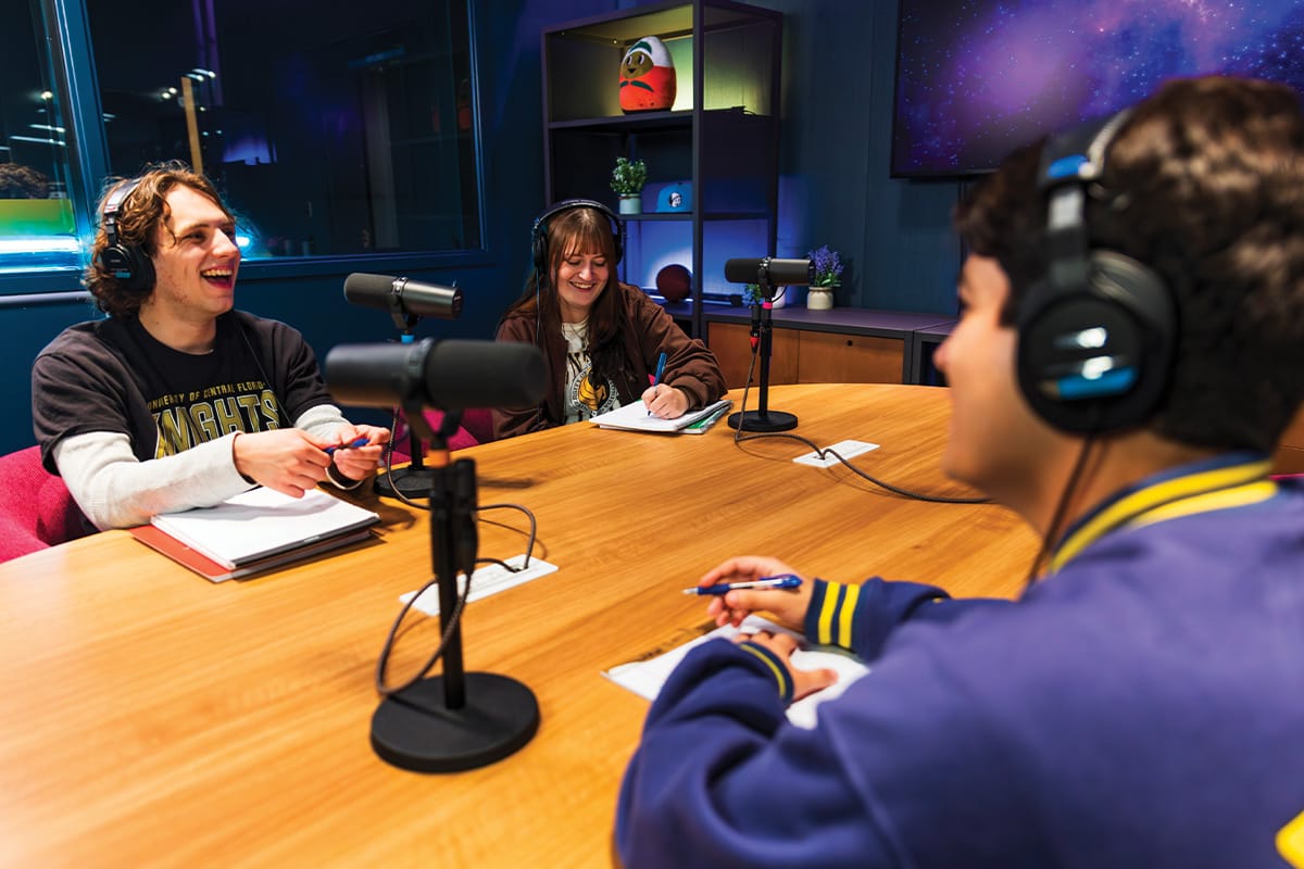 Two male college students and one female student, all wearing black headphones and laughing, sit at wooden table with black microphones on stands in front of each of them.