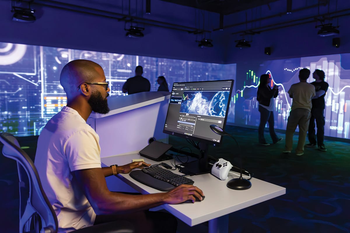 Side profile view of bearded man wearing glasses, who is seated at a gray rectangular table with computer monitor, keyboard, mouse and white game controller as surrounding walls display graphs and data sets to five people standing in the room.