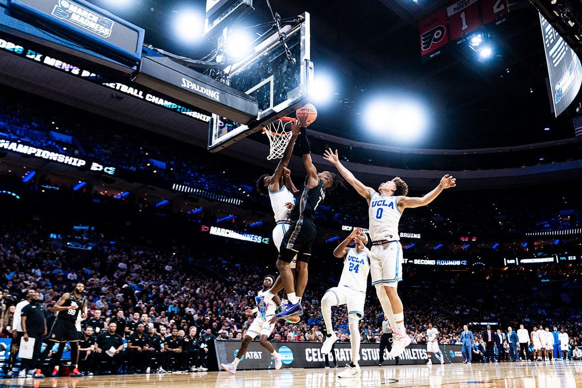 Basketball guard #1 Themus Fulks, in black UCF uniform, attempts a layup with three UCLA defenders circling him in crowded arena.