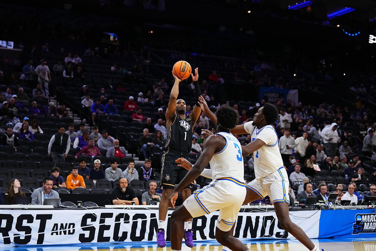 Basketball guard #1 Themus Fulks, in black UCF uniform, attempts a jump shot as two UCLA defenders close in with stands of people behind them watching.