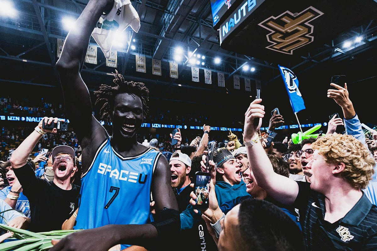 UCF center #7 Jon Bol, in blue SpaceU jersey, raises his right arm in celebration with crowd of students around him at UCF's Addition Financial Arena.