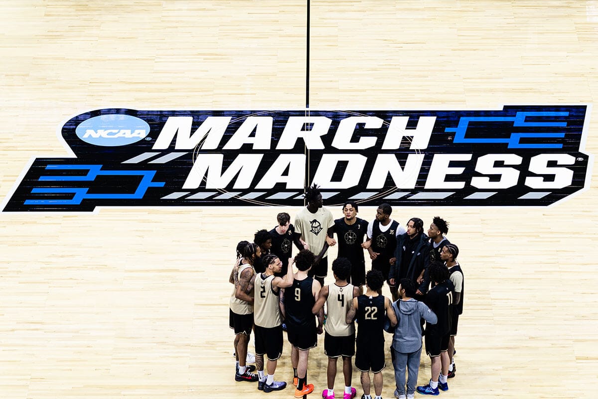 Aerial view of UCF players circled together in a huddle, standing underneath March Madness logo at center court.