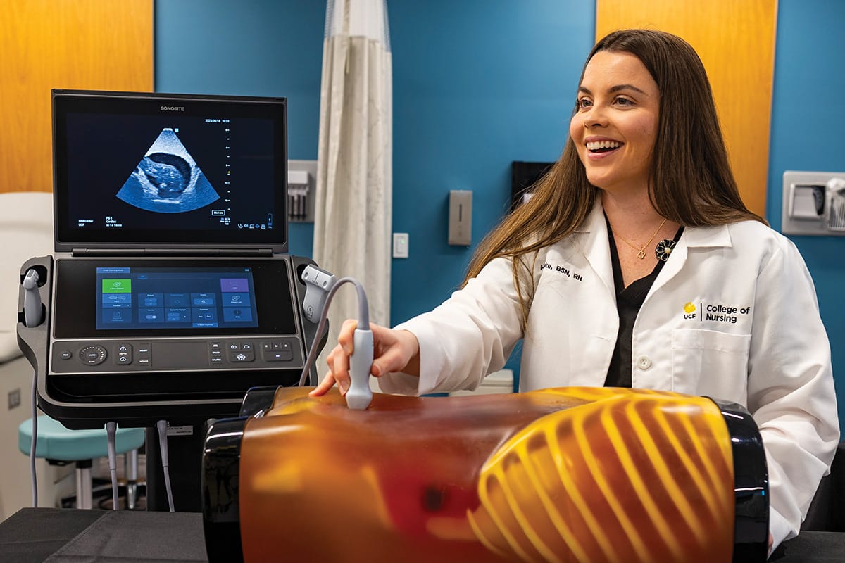 Brunette woman wearing white College of Nursing lab coat holds gray ultrasound tool to belly button of orange, translucent mannequin torso on black table in front of her in a medical room with blue walls, white divider curtain and monitor.