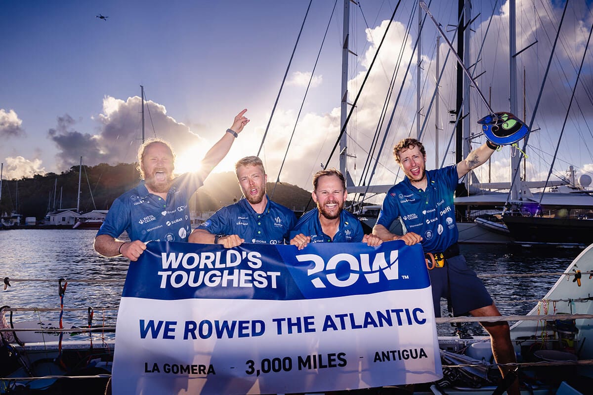 Four men in blue collared shirts stand on boat next to rippling water while holding a white and blue sign that reads: "World's Toughest Row. We rowed the Atlantic. La Gomera - 3,000 miles - Antigua" as sun rises behind a mountain in the background.