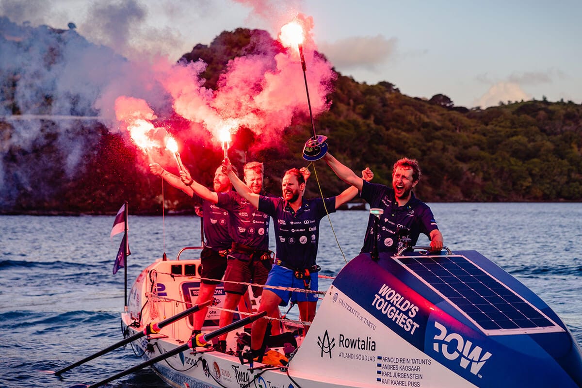 Four men wearing blue collared shirts stand on white and blue World's Toughest Row boat while holding red flairs as it drifts in rippling water with mountains in the background.