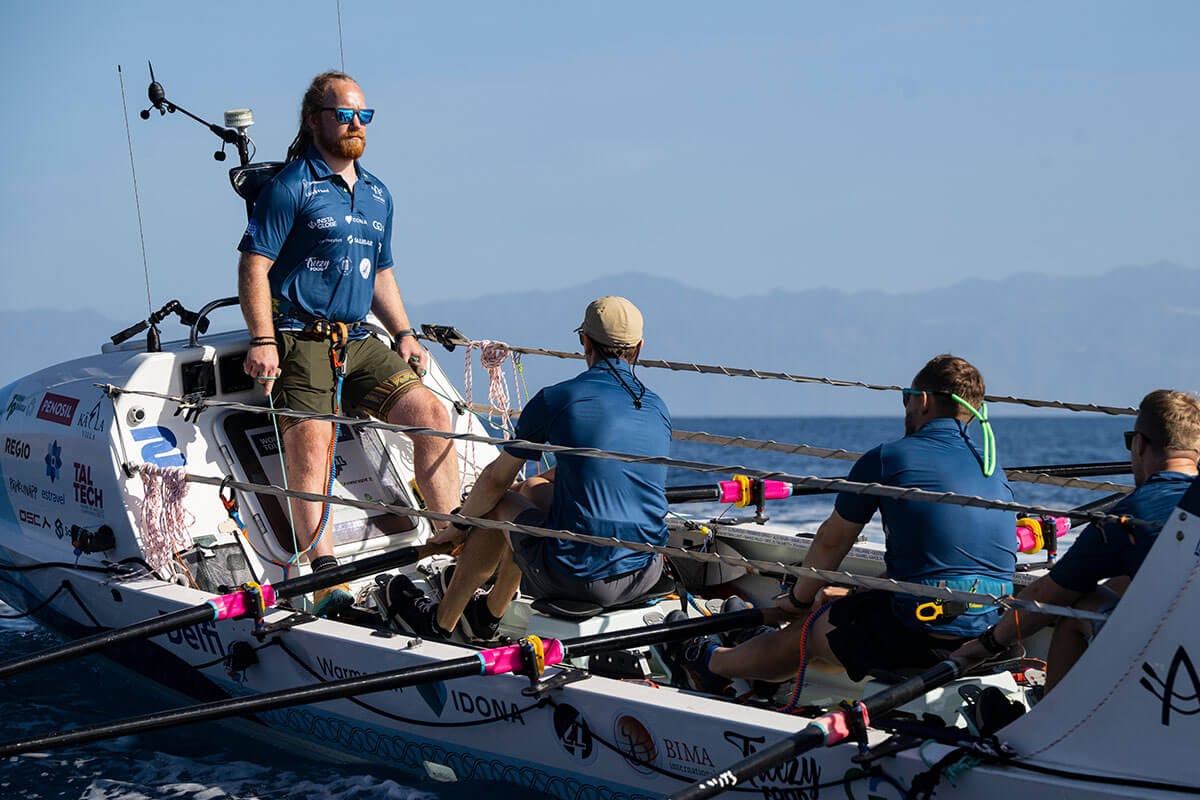 Bearded man wearing sunglasses in blue collared shirt and green cargo shorts stands at bow of white row boat as two men sit with oars in their hands, looking up at him.