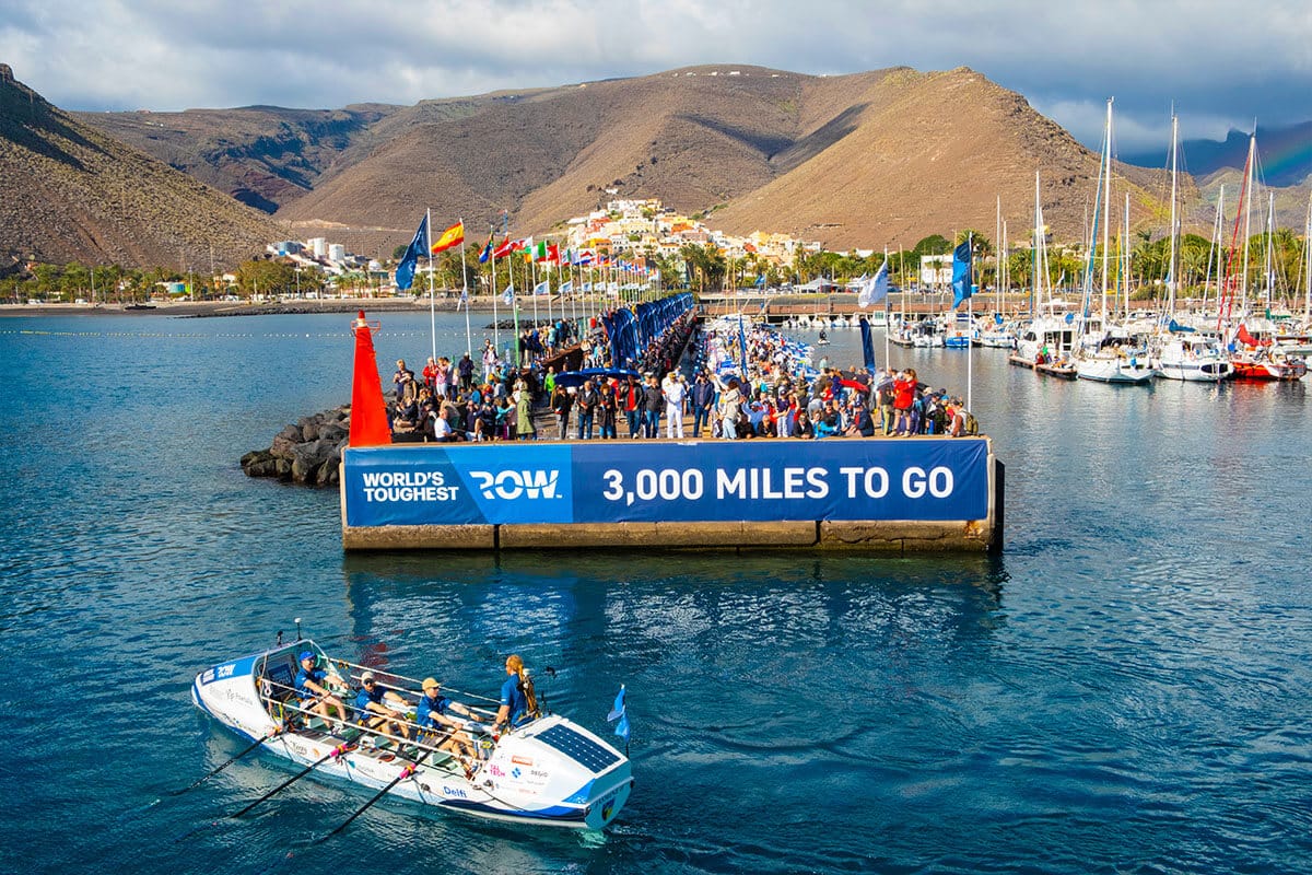 Aerial view of white row boat with three seated men and one man standing as it drafts away from large crowd of people gathered on dock decorated with flags of various countries and blue banner sign that reads: "World's Toughest Row. 3,000 Miles to Go."