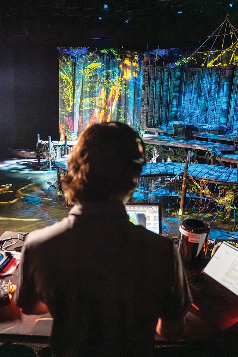 Man in suit jacket wearing headset stands in front of computer as he looks ahead at decorative stage setting, lit in blue, yellow and green hues.