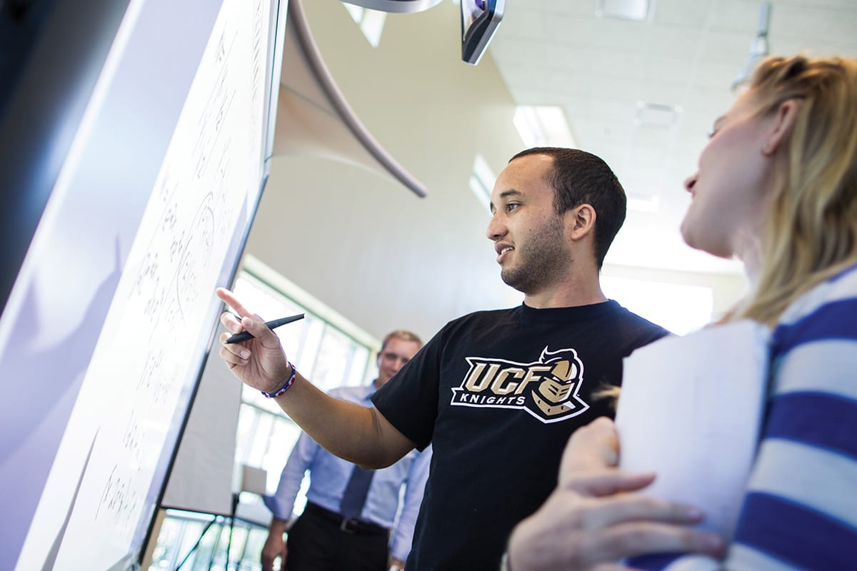 Man with dark hair and bearded stubble wearing black UCF shirt points to white screen with pen in his hand as young blonde woman in white and blue striped shirt observes next to him.