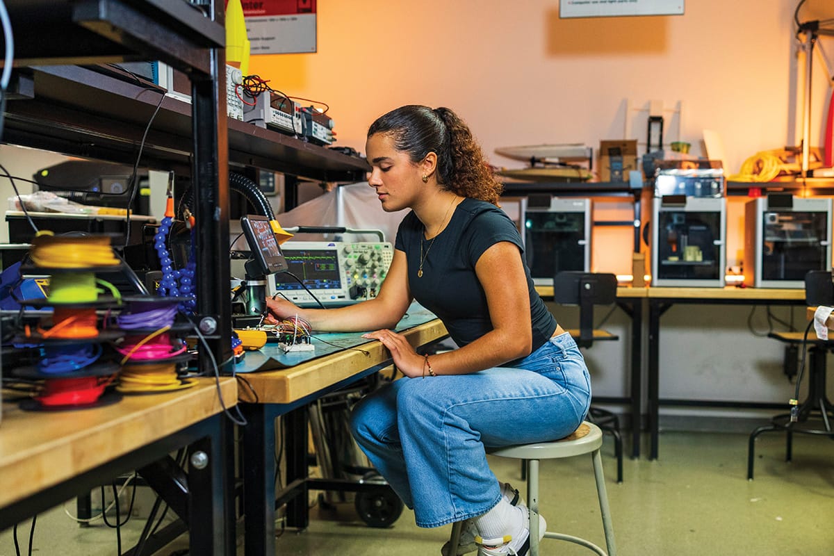 Young brunette woman with hair in ponytail, wearing black shirt, jeans and white sneakers, sits on metal stool at a wooden bench with many devices stacked on top.