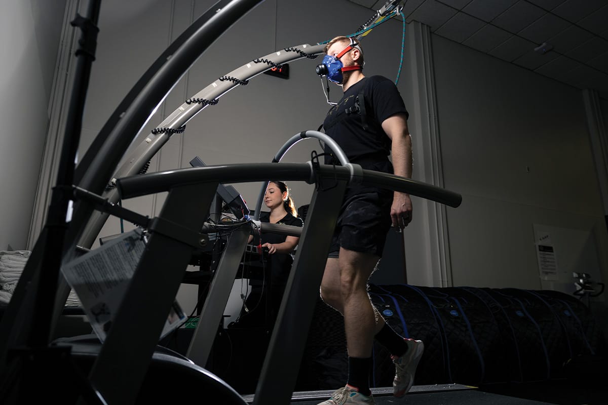 Man in black shirt, black shorts and white sneakers, wears a blue and red breathing mask as he walks on black treadmill with woman observing from his right side.
