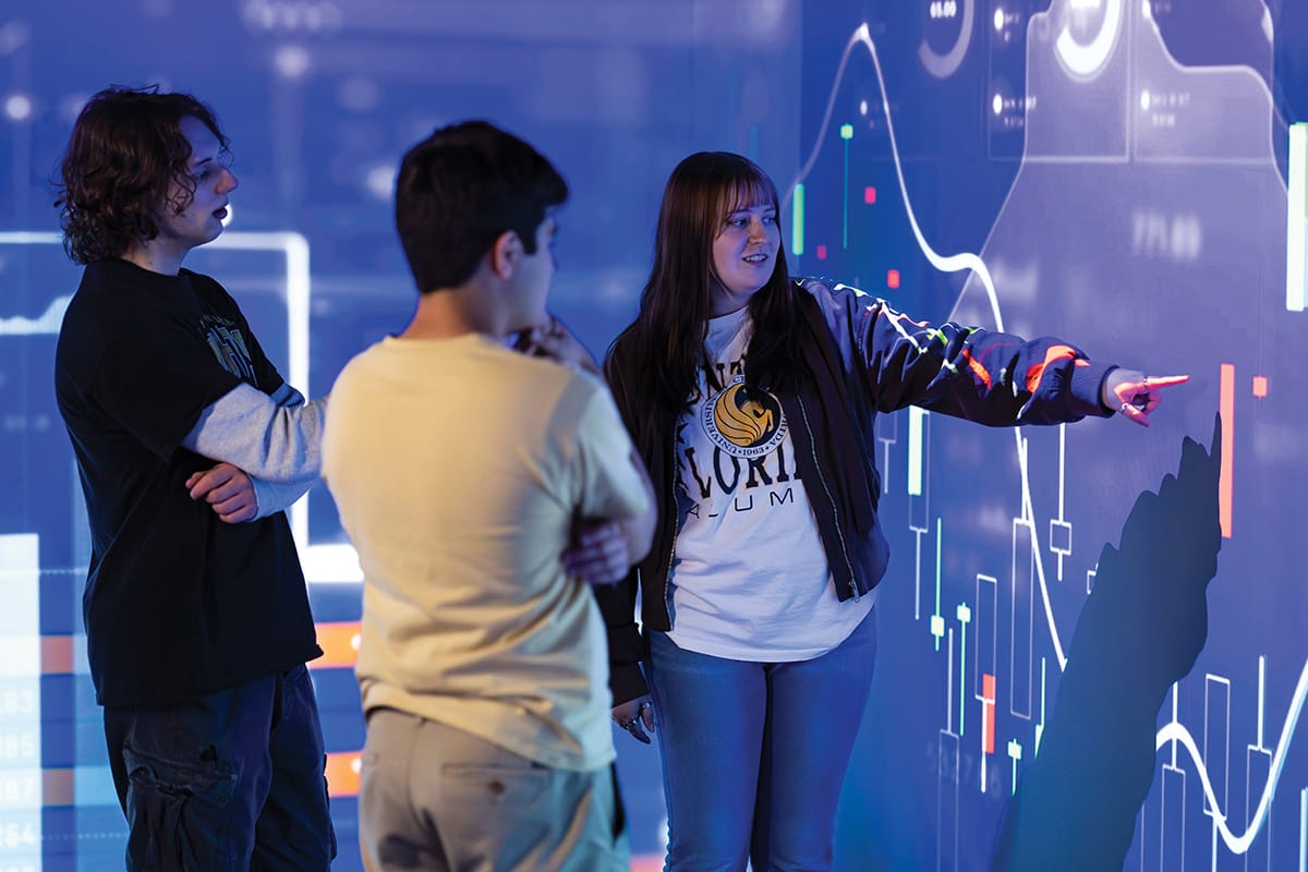 Brunette woman with bangs wearing dark jacket and white UCF T shirt points to projection screen on wall with graphing images as man in yellow T shirt observes.