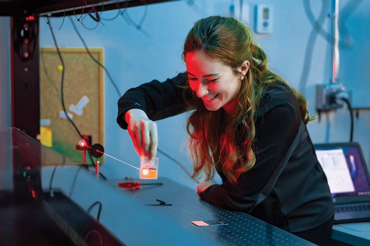 Woman with long wavy hair in black long sleeve shirt holds card in front of thin red laser beam in lab setting.