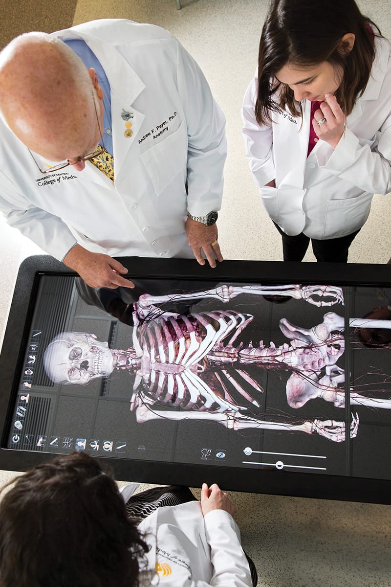 Aerial view of a bald man in white lab coat and brunette woman in white lab coat, staring down at rectangular black screen with skeleton projected onto it.