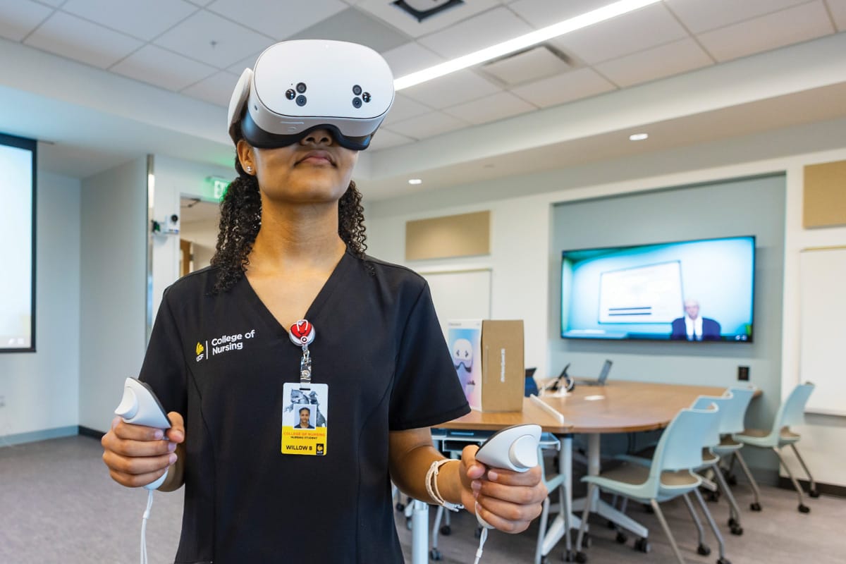 Woman in black nursing scrubs with UCF name badge clipped to her collar wears a VR headset while gripping two white controls in her hands in lab setting.