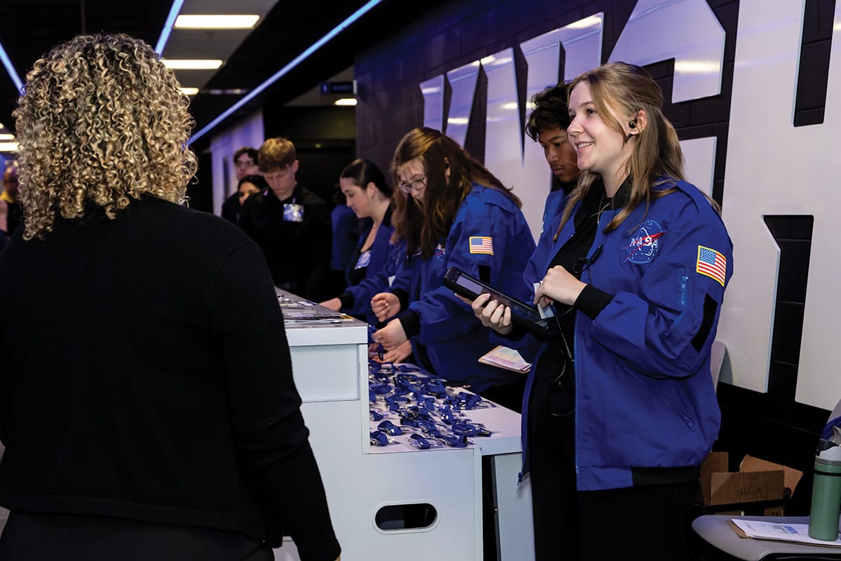 Six college students in blue NASA jackets stand behind check in counter and greet people.