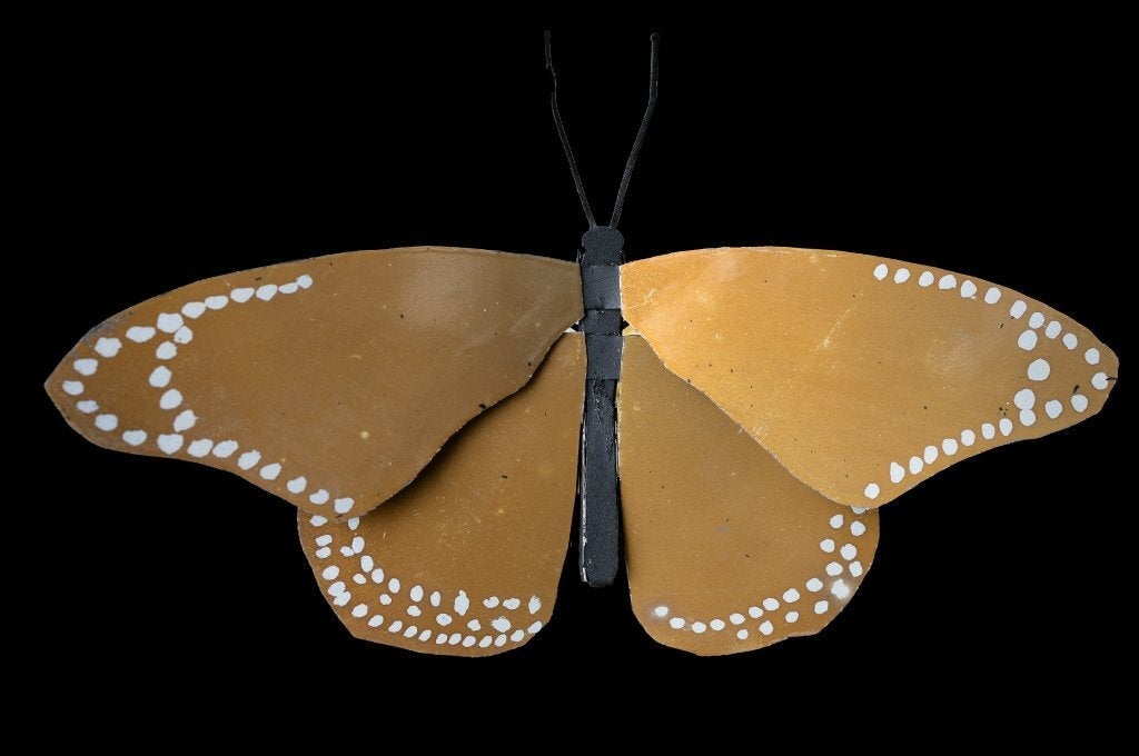 A handcrafted butterfly with warm amber wings dotted with white spots, photographed against a black background — illustrating structural color in nature.